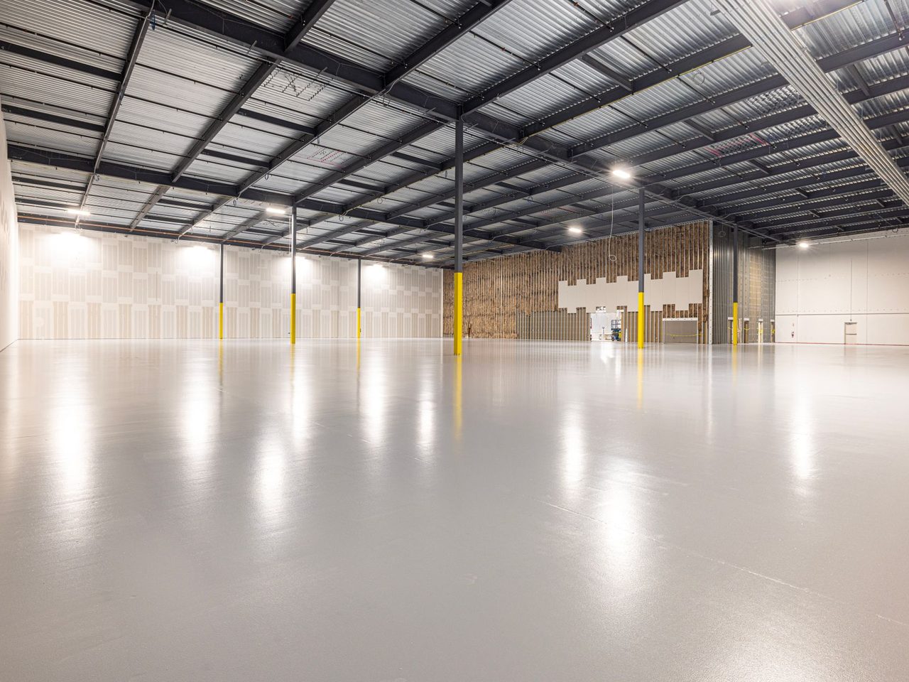 Large empty warehouse interior with glossy gray concrete floor, exposed steel roof trusses, overhead lights and yellow posts near the walls.