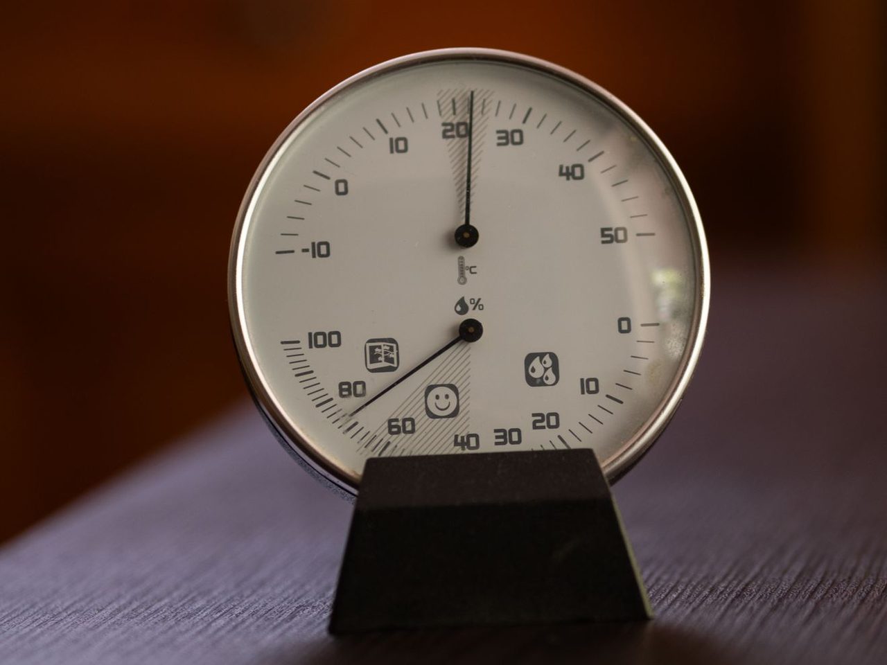 Round analog hygrometer and thermometer with white face and black needles mounted on a black stand against a dark brown background.