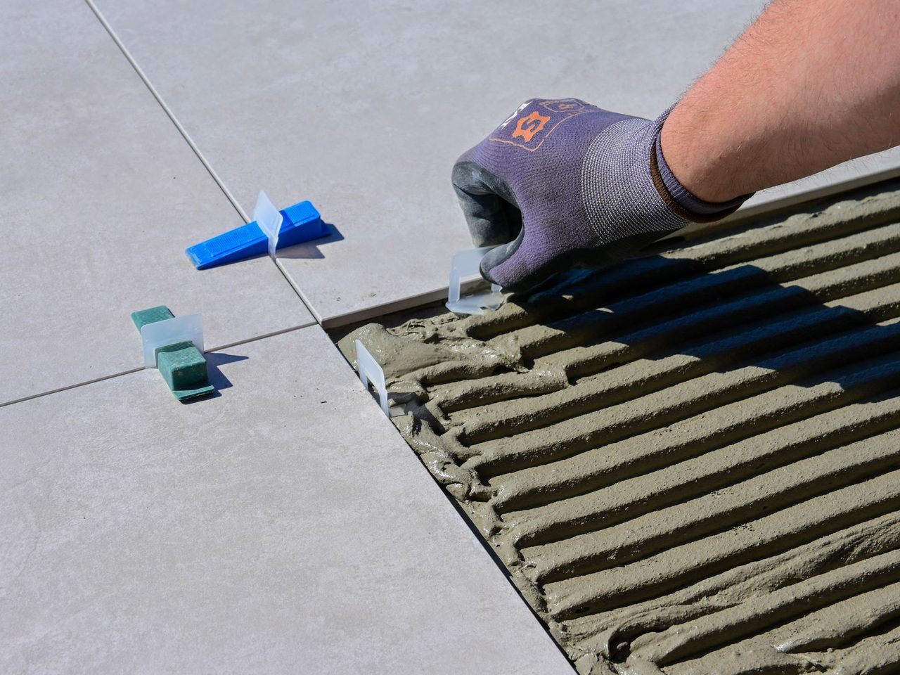 Gloved hand placing leveling clips and wedges between large floor tiles over notched thinset