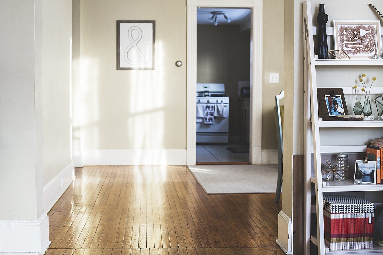 Sunlit hallway with an oak strip floor leading to a kitchen doorway, with a bookshelf at right