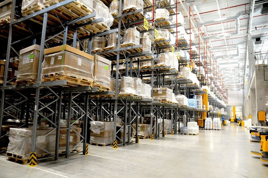 Bright modern warehouse with grey pallet racking holding stretch-wrapped pallets, yellow reach trucks along the aisle
