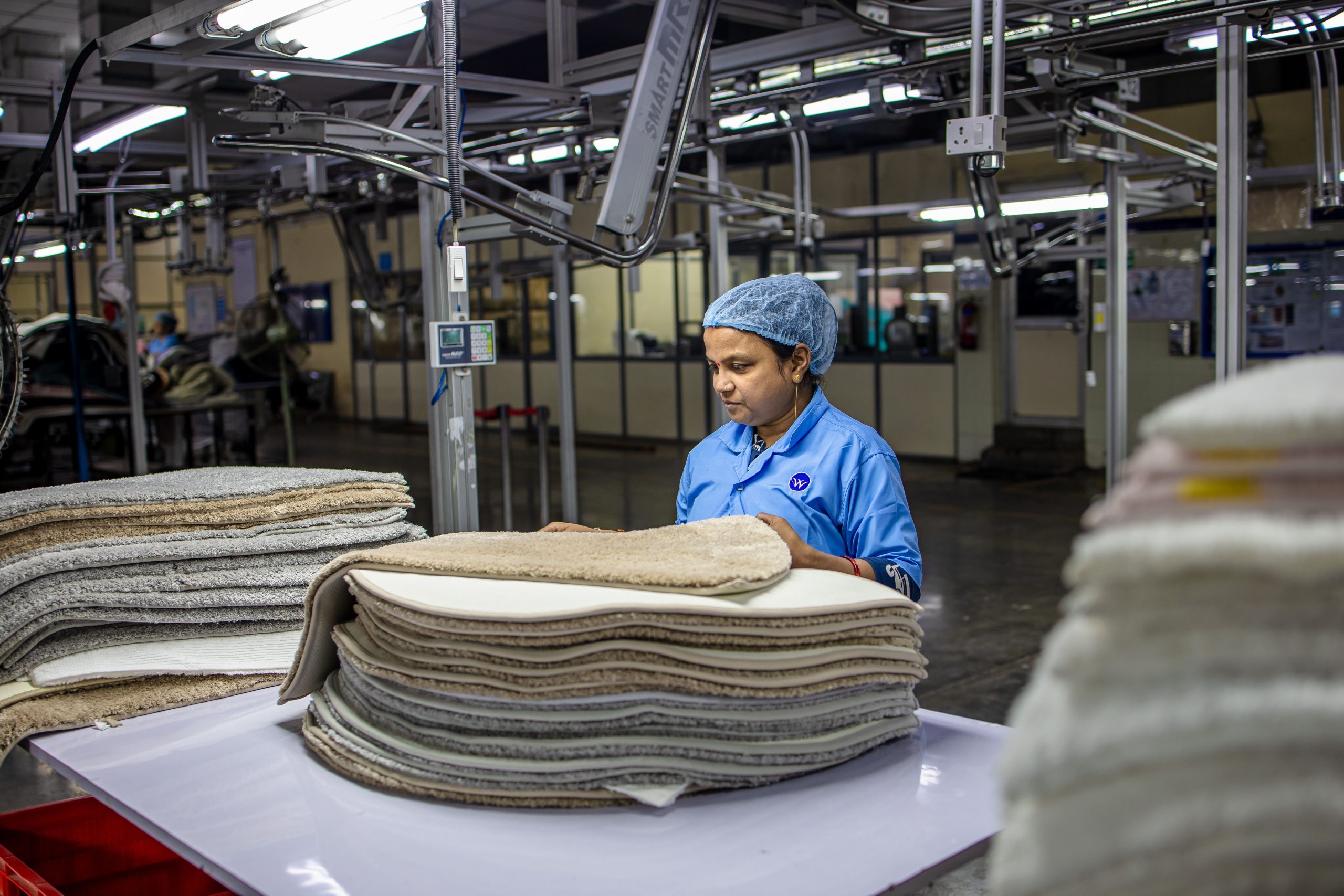 A textile worker in a factory setting inspects stacks of carpets under bright lights.
