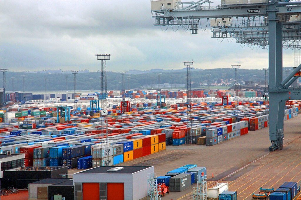 Container terminal yard with long rows of stacked shipping containers under a gantry crane on a cloudy day