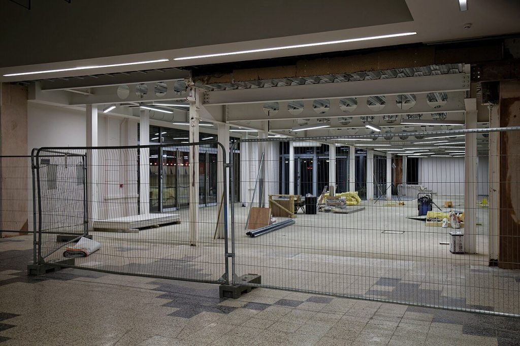 Interior fit-out behind temporary mesh fencing, with a bare floor, stacked materials and exposed ceiling beams