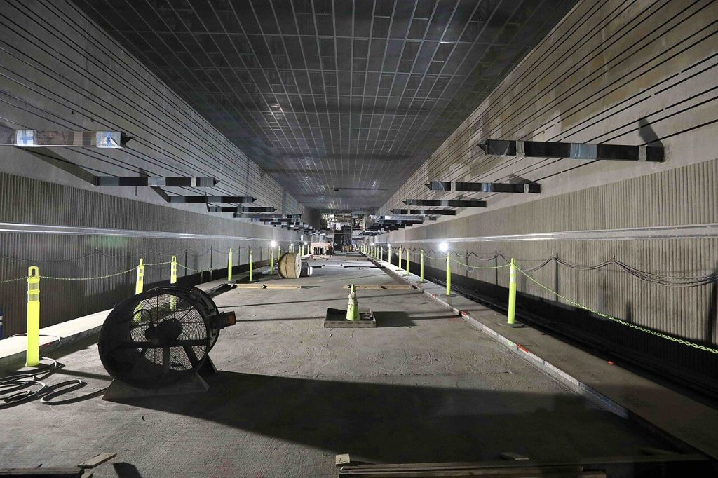 Long underground station interior under construction with a bare concrete floor, yellow bollards and a large fan