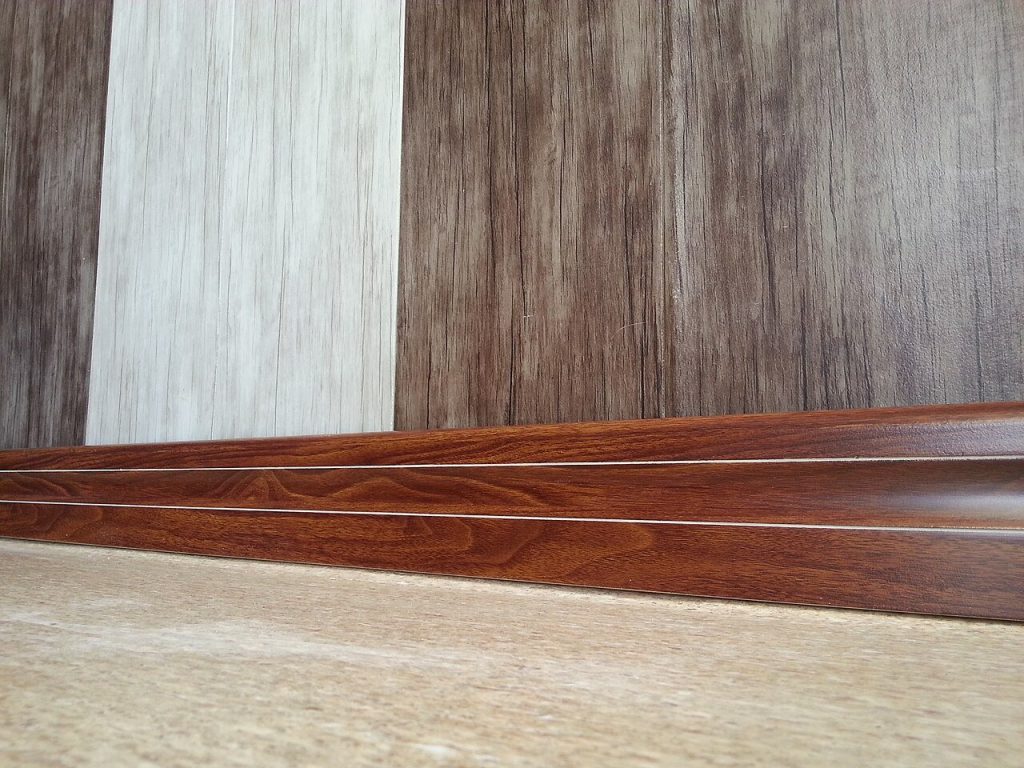 Wood-grain skirting profile on a floor in front of three wood-look decor samples in grey, cream and brown