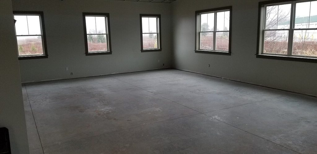Empty room with a bare concrete floor divided by saw-cut control joints, windows along two walls