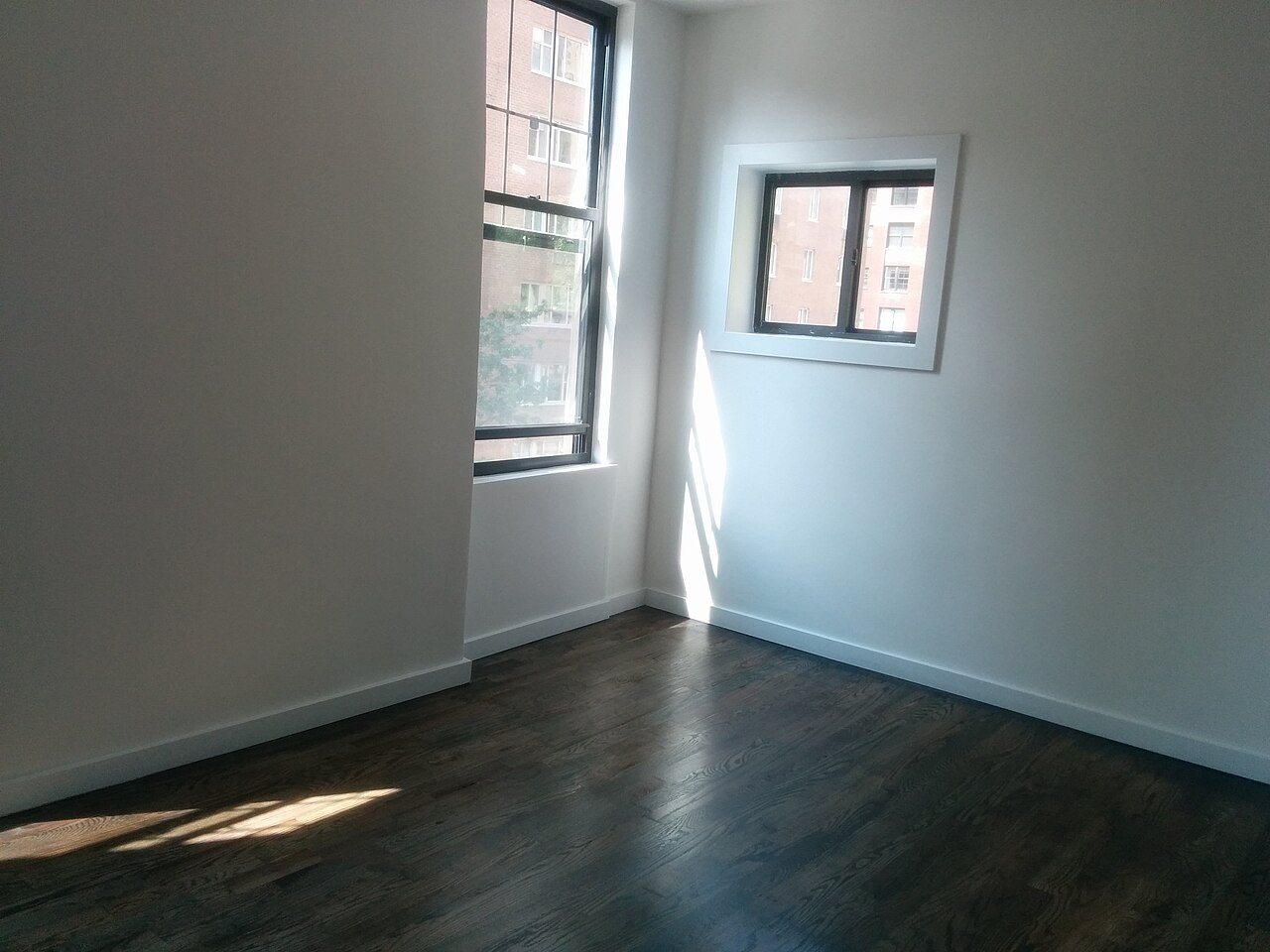 Empty apartment room with a dark stained oak strip floor, white walls and sunlight from two windows